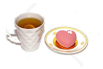 A pink heart-shaped cake on top is decorated with pearl beads on a saucer. next to a cup of tea and lemon