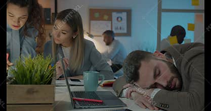 Young businessman sleeping at desk while colleagues working in dark office at night