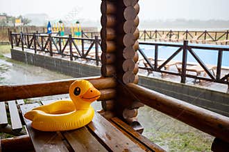 An inflatable duck lies on wooden table in gazebo at recreation center against the backdrop of pool covered with rain