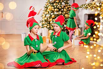Two cheerful happy girls-friends in costumes of Santa`s elves on the background of a Christmas tree and other children