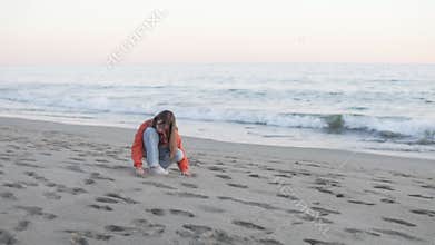 A brooding teenage girl sits on the sand by the sea in the evening