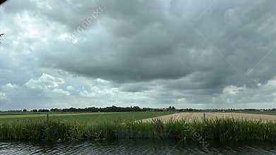Wide rural landscape with dark storm clouds over green fields, reed-lined