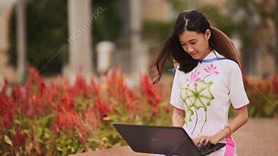 Pretty Asian girl sitting with a laptop in the park in national dress Ao Dai with a cap on the back of Non La