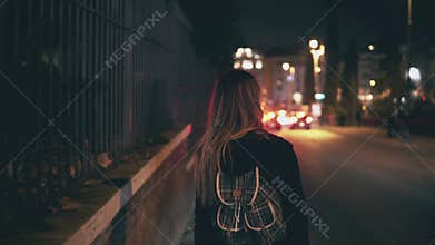 Back view of brunette woman walking near the road at the traffic time. Girl goes through the city late at night alone.