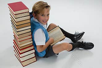 Schoolboy reading book while sitting against books stack on white background