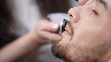 Close up of young bearded man getting haircut on his face by hairdresser at stylish barbershop. Image of female hands