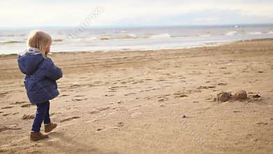 A little boy with blond hair walking along a sandy beach near the sea
