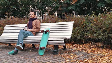 Young man with knitted scarf with skateboard sitting on bench in autumn park drinks coffee out of paper cup