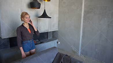 Female blonde speaks on cellular and walks around living room, smiling and laughing, sits on back of sofa.