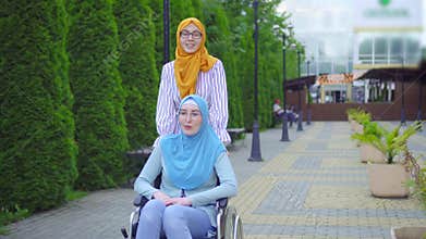 Young muslim disabled in a traditional scarf in a wheelchair communicates with a muslim friend on the street