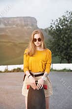 Beautiful and fashionable young woman in sunglasses and with a tattoo poses with a skateboard or longboard against the