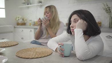Close-up of adult brunette Caucasian woman siting with coffee cup at the table. Her blond female friend drinking hot