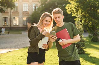 Two happy students checking smart phone content standing in the college park holding books and notebooks. Couple of cheerful
