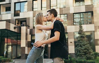 Loving young couple in stylish casual clothes kissing outdoors on the background of a modern building. Guy kisses a girl on a walk