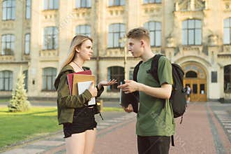 Two beautiful students guy and girl talking on background of university building, holding books, exercise books and laptop. A