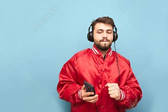 Funny man in a red jacket and a smartphone in his hand listening to music on headphones on a blue background. Bearded man in the