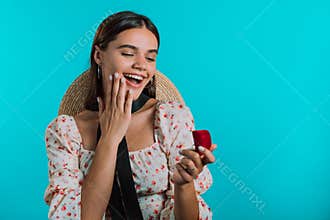 Attractive young woman holding small jewelry box with proposal diamond ring on blue wall background. Lady smiling, she