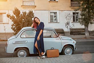 Travel time and new places idea, copy space. Woman in blue dress and suitcase on ground. Old vintage car on background
