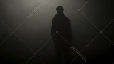 Black silhouette of man standing in smoky dark. Stock footage. Mysterious silhouette of young man stands in darkness lit