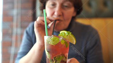 Adult woman drinking cocktail in cafe. Adult female sipping fresh fruit beverage through straw while sitting at table