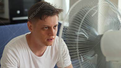 Overheated exhausted young man suffering from heat high temperature at home, sitting on sofa in front of ventilator