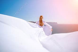 A blonde woman with long hair stands on a sunny seashore in a flowing white dress, with the silk fabric waving in the