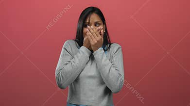 Woman with long dark hair in gray sweater covers mouth in surprise against red wall background illustrating shock emotion and