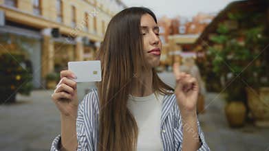 Young hispanic woman holds white creditcard and points index finger in refusal gesture on urban street; rejection