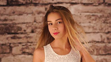 Young teenage girl is staying near the wall, shaking her head, playing with hair and looking at the camera. The wind is