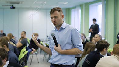 Professional coach in blue shirt speaking into the microphone and gesticulating at workshop for future top managers of a