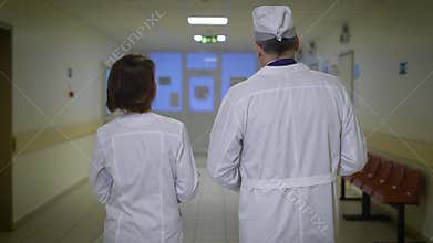 Back view of male and female doctors dressed in lab coats walking through the corridor of the ward and talking to each