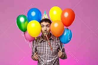 Young man celebrating birthday, holding colorful baloons over purple background.