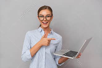 Cheerful happy business young woman posing isolated over grey wall background using laptop computer pointing.