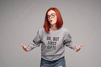 Swag and confident red haired girl in jeans and sweatshirt posing at gray studio.