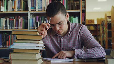 Young handsome teenage man wearing glasses sitting at table in university library thinking over calculations in copybook