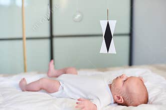 Adorable baby boy in white sunny bedroom lying and looks at Munari Montessori mobile.