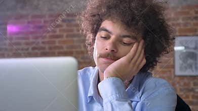 Bored nerdy office worker typing on laptop and sitting in modern office, trying not to sleep from boredom