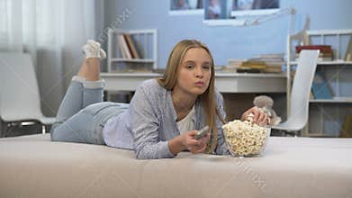 Teenage girl watching tv in room with remote control in hand and eating pop corn