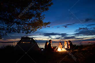 Night summer camping on shore. Group of young tourists around campfire near tent under evening sky