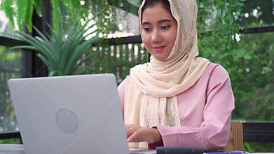 Beautiful young smiling asian muslim woman working on laptop sitting in living room at home.