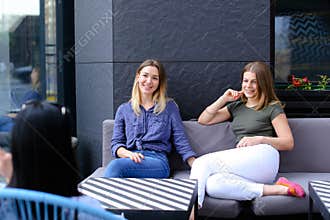 Beautiful female friends talking at cafe and sitting on sofa near window with flowers.