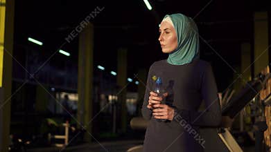 Young sporty muslim girl in hijab drinking water from bottle after workout