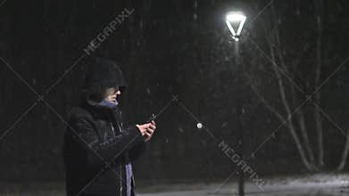 The man in the street light. The guy looks into his smartphone. It`s snowing against the background of lamp. Slow motion