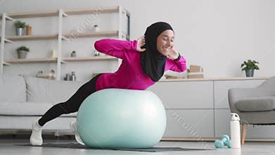 Back and core strengthening workout. Young muslim woman wearing hijab doing abs exercises on fitness ball at home