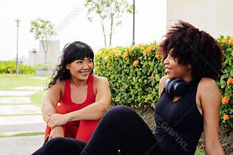 Diverse group of two female fitness friends taking break and talking in the park. Young women resting and smiling