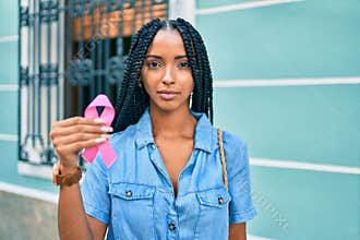 Young african american woman with relaxed expression holding pink breast cancer ribbon at the city
