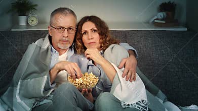 A middle-aged couple watches horror movies on TV and eats popcorn on the couch.