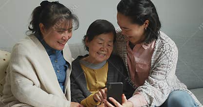 Smiling senior asian woman using smartphone with adult daughter and granddaughter embracing
