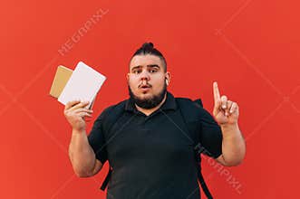 Surprised fat young male student with beard stands on red background with book and notebook in hand and shows finger upward on