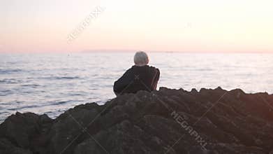 Handsome blond boy sitting on rocks by the sea at sunset,